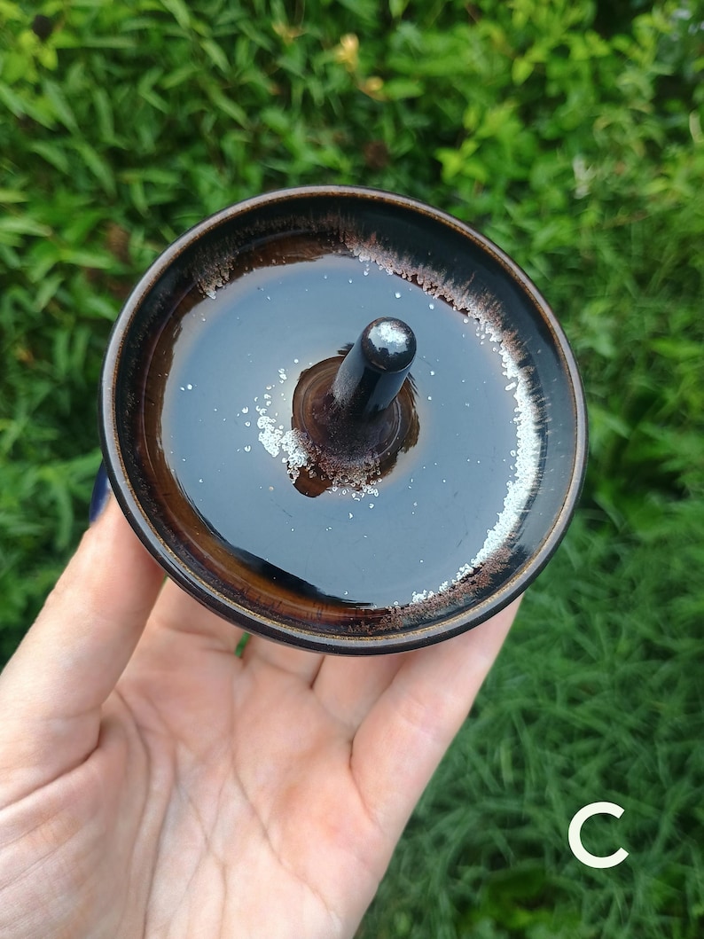 Puede incluir: Un cuenco de cer&aacute;mica circular para lana con un poste central para sujetar el hilo. El cuenco presenta un esmalte marr&oacute;n oscuro brillante con un interior marr&oacute;n m&aacute;s claro y un borde moteado blanco distintivo. La letra C es visible abajo a la derecha.