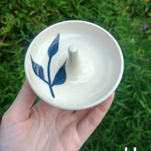 Puede incluir: Un plato de cer&aacute;mica redondo de color blanco roto con un poste c&oacute;nico central para guardar anillos. La superficie interior est&aacute; decorada con un motivo bot&aacute;nico de hojas de color azul oscuro pintado a mano. Tiene un acabado liso y brillante.