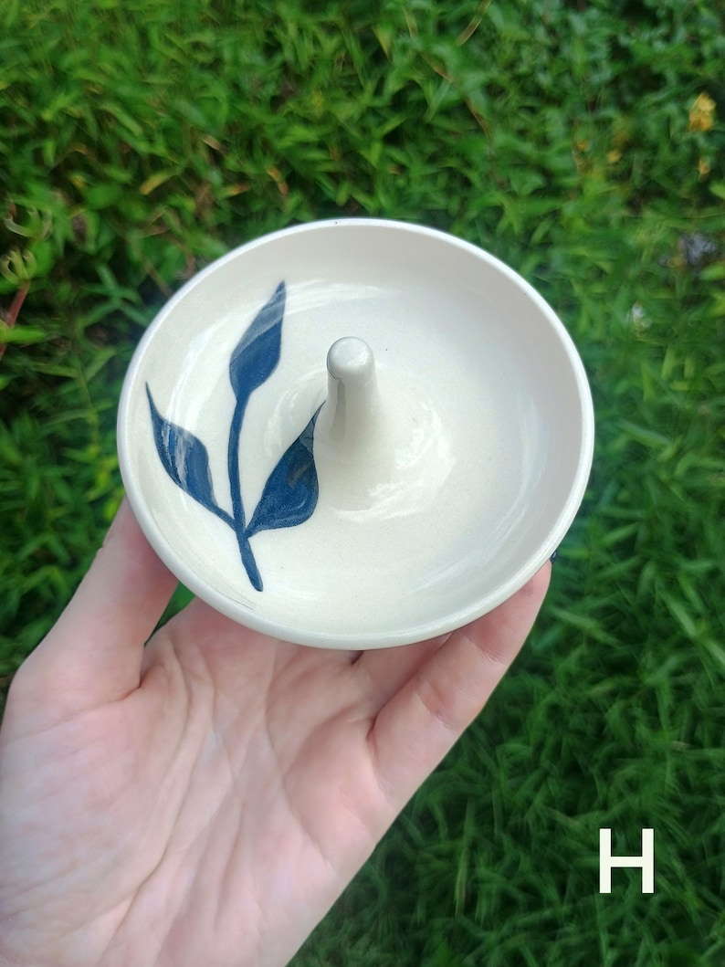 Puede incluir: Un plato de cer&aacute;mica redondo de color blanco roto con un poste c&oacute;nico central para guardar anillos. La superficie interior est&aacute; decorada con un motivo bot&aacute;nico de hojas de color azul oscuro pintado a mano. Tiene un acabado liso y brillante.