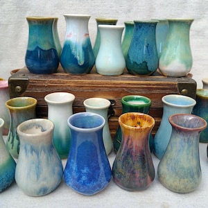 May include: A collection of small, handcrafted ceramic vases in various colors, including blue, green, and white. Each vase has a unique glaze, with some featuring a two-tone effect. The vases are displayed on a wooden surface.