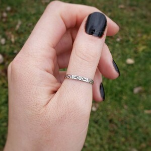 May include: A silver ring with a delicate floral design on the band. The ring is worn on the thumb of a hand with black nail polish.