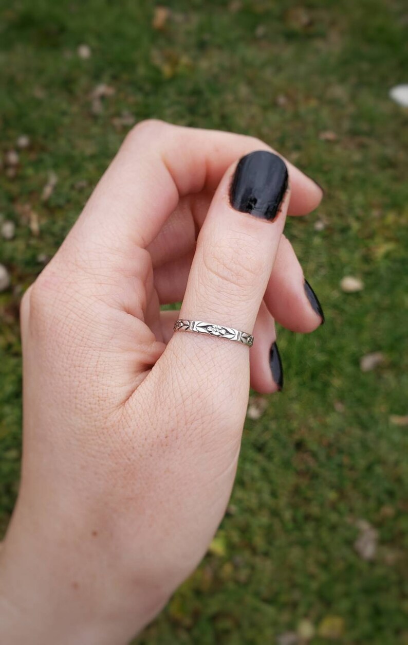 May include: A silver ring with a delicate floral design on the band. The ring is worn on the thumb of a hand with black nail polish.