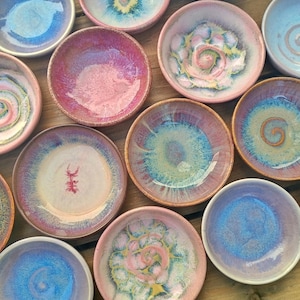 May include: A collection of small, round ceramic bowls featuring a variety of reactive glazes in shades of pink, blue, purple, and white. The bowls display diverse surface patterns, including central spirals, mottled textures, and smooth gradients.