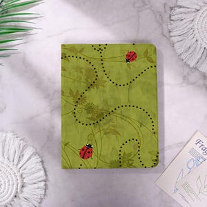 May include: A rectangular notebook with a light green cover featuring a subtle leaf pattern, two red ladybugs with black spots, and curved dotted lines that trace paths across the surface. The design includes delicate dark green vine illustrations.