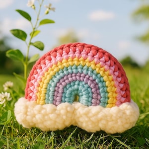 Puede incluir: Un juguete de arcoíris tejido a ganchillo con una base de nube blanca y esponjosa. El arcoíris presenta bandas de rojo, rosa, amarillo, verde, azul y morado. El juguete está sobre hierba verde con cielo azul.