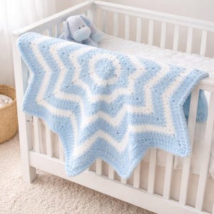 Könnte beinhalten: Eine weiche, blau-weiß sternförmige Babydecke, die über einem weißen Kinderbett liegt. Die Decke hat ein großes Sterndesign mit abwechselnd blauen und weißen Abschnitten. Ein blaues Stofftier befindet sich im Kinderbett.