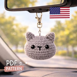 May include: A crocheted gray cat head keychain with black button eyes and a black 'T' mouth hangs from a gold-colored clasp. The keychain is suspended from a car's rearview mirror. The image includes a small American flag and the text 'PDF PATTERN'.