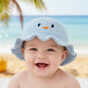 May include: A light blue crochet baby hat with a bird face design, featuring black eyes and an orange beak. The hat has a scalloped brim and is worn by a smiling baby. The background shows a beach scene.