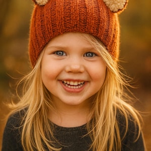 May include: A child wearing a rust-colored knit hat with bear ears and a gray long-sleeved shirt. The hat has cream-colored pom-poms on the ears. The child is smiling with blonde hair and blue eyes.