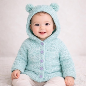 May include: A baby wearing a pale blue knitted hooded jacket with bear ears and purple buttons. The jacket is fastened and the baby is smiling. The baby is wearing light-coloured trousers.