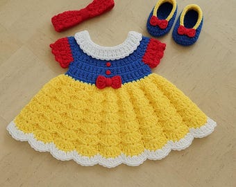 May include: A crocheted Snow White costume for a baby, featuring a yellow and white dress, blue bodice, red bow, and matching headband and shoes.