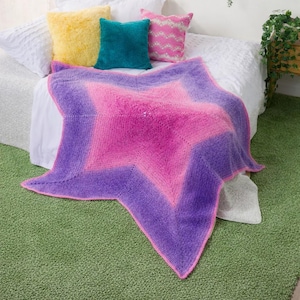 May include: A star-shaped blanket in pink and purple hues, edged with pink, is draped over a bed. Cushions in yellow, teal, and pink chevron patterns are arranged around it. The bed is covered with a white duvet.