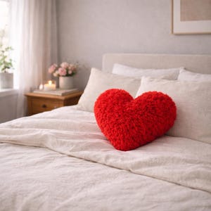 May include: A plush, red heart-shaped pillow rests on a bed with white linens. The pillow is the focal point, with a textured, fuzzy surface. The bedroom setting includes a nightstand, flowers, and soft lighting, creating a cozy atmosphere.