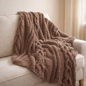 May include: A brown cable-knit throw blanket draped over a white sofa. The blanket features a thick, textured weave with prominent cable patterns along the edges. The warm, neutral color and cozy texture suggest comfort and relaxation, perfect for home decor.