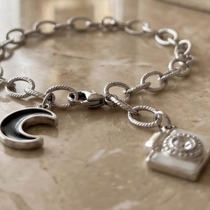 Peut inclure: Un bracelet à maillons argentés doté de deux breloques pendantes. L'une est un croissant de lune avec un émail noir, et l'autre est un appareil photo carré avec une pierre centrale transparente et des détails en émail blanc.