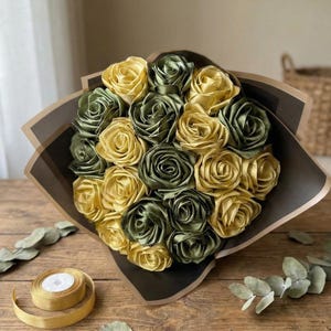 Handmade forever flower bouquet featuring olive green and golden yellow satin ribbon roses wrapped in elegant translucent brown paper. A luxury long lasting floral gift designed for weddings, anniversaries, Mother’s Day, and romantic occasions. Displayed in a modern neutral interior setting with jewelry box and soft natural light. A timeless artificial rose bouquet that never fades and serves as a keepsake floral arrangement.
