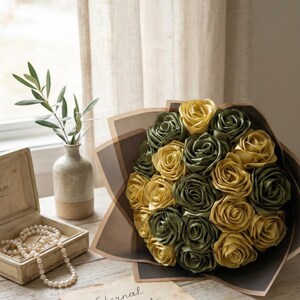 Handmade forever flower bouquet featuring olive green and golden yellow satin ribbon roses wrapped in elegant translucent brown paper. A luxury long lasting floral gift designed for weddings, anniversaries, Mother’s Day, and romantic occasions. Displayed in a modern neutral interior setting with jewelry box and soft natural light. A timeless artificial rose bouquet that never fades and serves as a keepsake floral arrangement.