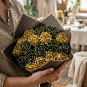 Handmade forever flower bouquet featuring olive green and golden yellow satin ribbon roses wrapped in elegant translucent brown paper. A luxury long lasting floral gift designed for weddings, anniversaries, Mother’s Day, and romantic occasions. Displayed in a modern neutral interior setting with jewelry box and soft natural light. A timeless artificial rose bouquet that never fades and serves as a keepsake floral arrangement.