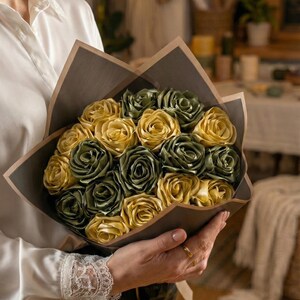 Handmade forever flower bouquet featuring olive green and golden yellow satin ribbon roses wrapped in elegant translucent brown paper. A luxury long lasting floral gift designed for weddings, anniversaries, Mother’s Day, and romantic occasions. Displayed in a modern neutral interior setting with jewelry box and soft natural light. A timeless artificial rose bouquet that never fades and serves as a keepsake floral arrangement.