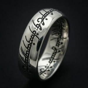 Könnte beinhalten: Eine weiße Keramik-Tasse mit schwarzem Henkel und dem Text "But First, Coffee" in schwarzer, handschriftlicher Schrift. Die Tasse steht auf einer weißen Oberfläche.