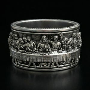 May include: A silver-colored ring with a detailed relief of the Last Supper scene. The ring features a detailed depiction of the figures around a table, with intricate detailing and a polished finish. The ring is a religious-themed accessory.