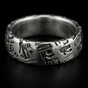 May include: A silver-colored ring with engraved hieroglyphic symbols. The ring has a wide band and a polished finish, with the symbols wrapping around the entire circumference. The ring is set against a black background.