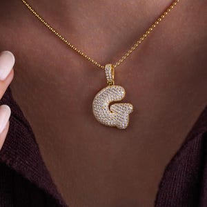 Puede incluir: Un collar dorado con un colgante en forma de la letra "G". El colgante está incrustado con pequeñas piedras blancas brillantes. El collar se muestra sobre un fondo oscuro.