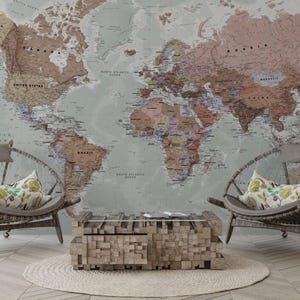 Puede incluir: Un gran mural de mapa del mundo en tonos apagados de marrón, beige y verde sirve de telón de fondo para una escena de sala de estar. Dos sillas con respaldo redondo con cojines grises y almohadas florales frente a una mesa de centro de madera. Una alfombra en el suelo.