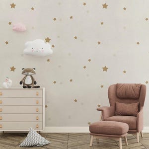 May include: A nursery scene with a light beige wall adorned with gold star decals. A white dresser with gold handles holds a fox plushie and a unicorn figurine. A pink cloud and a white cloud with a face are mounted on the wall. A pink armchair and ottoman complete the room.