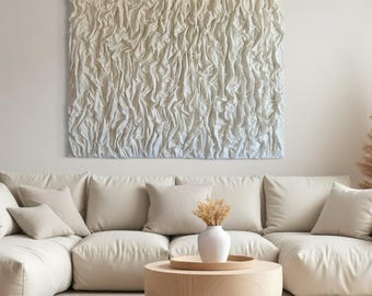 Könnte beinhalten: Ein großes, strukturiertes, cremefarbenes Wandkunstwerk hängt über einem cremefarbenen Ecksofa. Das Kunstwerk hat ein geriffeltes, stoffähnliches Aussehen. Ein runder Couchtisch aus Holz und eine weiße Vase mit getrockneten Blumen vervollständigen das Wohnzimmer.