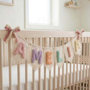 May include: A fabric banner with the name "AMELIA" in colorful, textured letters, strung across a crib. The banner features a cream-colored background with lace trim and is adorned with pink bows.