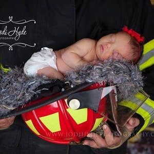 May include: A newborn baby wearing a red headband is sleeping in a firefighter's helmet. The helmet is red with a yellow stripe and is held by a person wearing a firefighter's uniform. The text "Jodi Hyde Photography" is visible in the top left corner of the image.