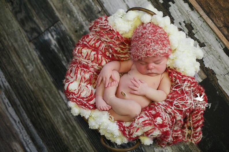 Peut inclure: Un nouveau-n&eacute; portant un bonnet tricot&eacute; rouge et blanc dort dans un panier tricot&eacute; blanc et rouge. Le panier est pos&eacute; sur un sol en bois.