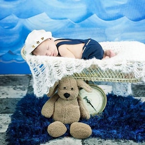 Peut inclure: Un nouveau-n&eacute; portant un chapeau de marin et une salopette bleue dort dans un panier en osier blanc. Un ours en peluche brun est assis sur un tapis bleu avec une horloge vintage.