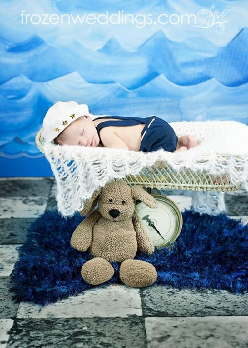 Peut inclure: Un nouveau-n&eacute; portant un chapeau de marin et une salopette bleue dort dans un panier en osier blanc. Un ours en peluche brun est assis sur un tapis bleu avec une horloge vintage.