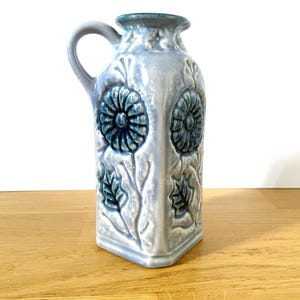 May include: A light blue ceramic pitcher with a handle, featuring embossed floral designs in teal. The vase has a square base and a slightly flared top, with a teal rim. The pitcher is approximately 8 inches tall.