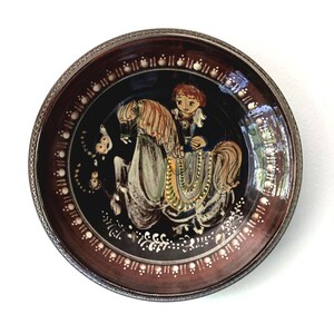 May include: A decorative ceramic plate with a dark brown rim and a black center. The plate features a hand-painted image of a person riding a horse. The rim has a textured design with white dots. The plate is a unique piece of art.