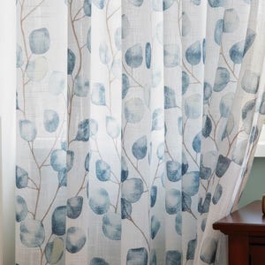 Könnte beinhalten: Weißer, transparenter Vorhang mit einem Aquarell-Eukalyptusblatt-Design in Blau- und Grautönen. Der Vorhang weist ein sich wiederholendes Muster aus Blättern und Zweigen auf, das eine natürliche und luftige Ästhetik erzeugt. Der Vorhang hängt vor einem Fenster.