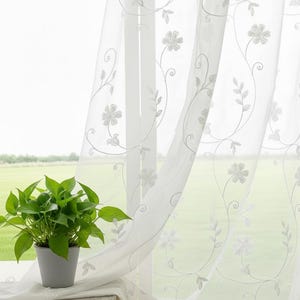 Puede incluir: Paneles de cortina blancos transparentes con patrones florales y de vid bordados. Las cortinas cuelgan frente a una ventana, con una planta en maceta en el alféizar. El fondo muestra un campo verde y un cielo brillante.