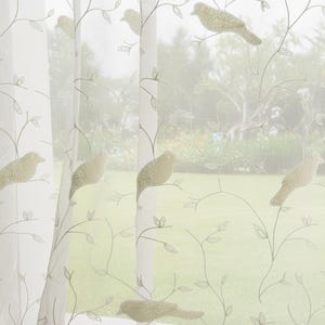 Puede incluir: Paneles de cortinas blancos transparentes con un delicado diseño de pájaros y ramas. Los pájaros son de color dorado apagado, y las ramas y hojas son de un verde suave. Las cortinas cuelgan frente a una ventana.