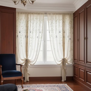 May include: Cream-coloured sheer curtains adorned with green leaves and bird motifs. The curtains are tied back, revealing a white window. The room includes dark wood furniture and a blue chair, creating a warm atmosphere.
