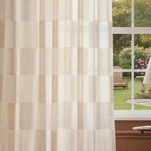 Può includere: Tenda trasparente color crema con un motivo geometrico a blocchi rettangolari. La tenda copre parzialmente una finestra che si affaccia su un giardino con una sedia e una lampada.