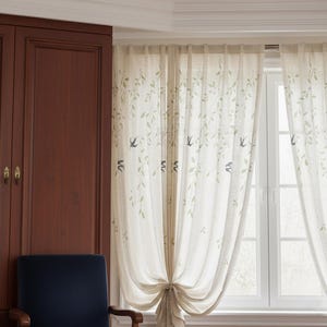 May include: Cream-coloured sheer curtains featuring a pattern of green leaves and blue birds. The curtains are tied back with a matching fabric tie, and hang in front of a white window. A dark wood cabinet and a blue chair are visible.