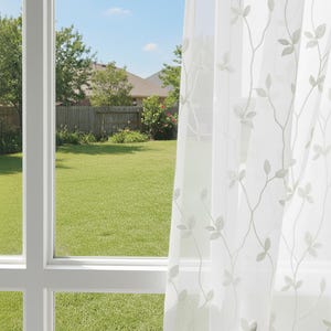 Könnte beinhalten: Transparenter weißer Vorhang mit einem zarten Blatt- und Rankenmuster. Der Vorhang verdeckt teilweise ein Fenster mit Blick auf einen grünen Rasen, Bäume und einen blauen Himmel. Der Fensterrahmen ist weiß.