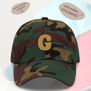 Könnte beinhalten: Tarnfarbene Baseballkappe mit einem großen goldenen "G" auf der Vorderseite. Die Kappe hat einen gebogenen Schirm und einen schwarzen Knopf oben. Weitere Kappen in Hellblau und Rosa sind im Hintergrund zu sehen.