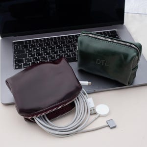 May include: Two leather pouches, one burgundy and one dark green, with silver zippers. The burgundy pouch is open, revealing a gray charging cable and a white Apple Watch charger. The green pouch has "DTL" embossed on it. A laptop is in the background.