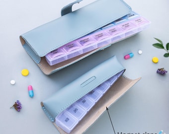 May include: A light blue leather pill box with a magnetic clasp. The pill box has a clear plastic insert with compartments for each day of the week and for morning, afternoon, and evening doses.