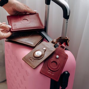 May include: A collection of leather travel accessories in shades of brown and burgundy. The items include wallets and luggage tags, some with a circular cutout and the initials "GLS" embossed. A pink suitcase is in the background.