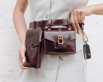 May include: A burgundy leather belt bag with two attached pouches and a gold-colored buckle. The belt is worn around the waist and has a matching burgundy leather strap. A key fob with keys is attached to the belt.