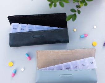 May include: Two pill cases, one black and one light blue, with a clear pill organizer inside. The cases are open and the pill organizer is visible. The pill organizer has days of the week labeled on it.